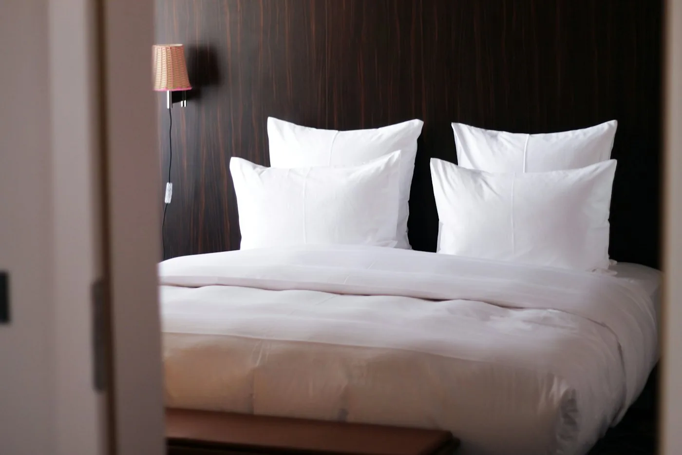 Hotel bed made up with crisp white linen and four large white pillows against a dark wood headboard